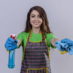 young beautiful woman with long wavy hair wearing apron and rubber gloves holding rug and cleaning spray looking confident and positive smiling ready to clean standing over white background
